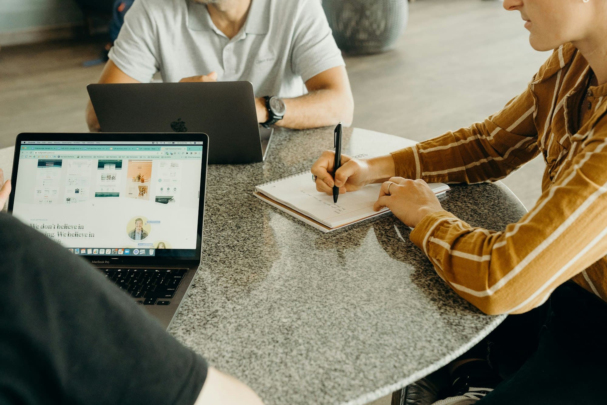 Trois personnes discutent d'un projet web autour d'une table, avec des ordinateurs et un carnet